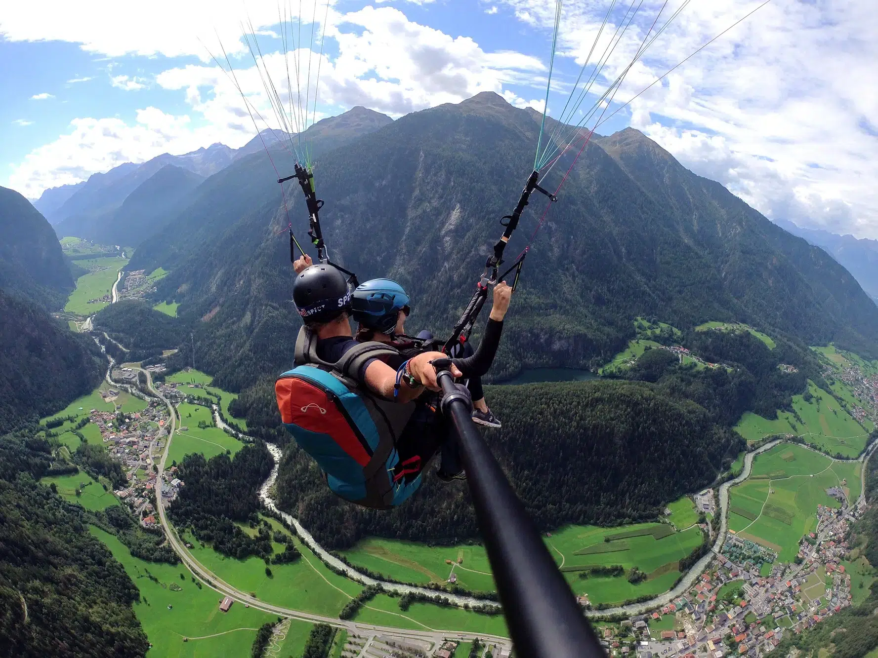 Zwei Personen beim Gleitschirmfliegen über dem grünen Ötztal mit seinen Bergen unter einem teilweise bewölkten Himmel.