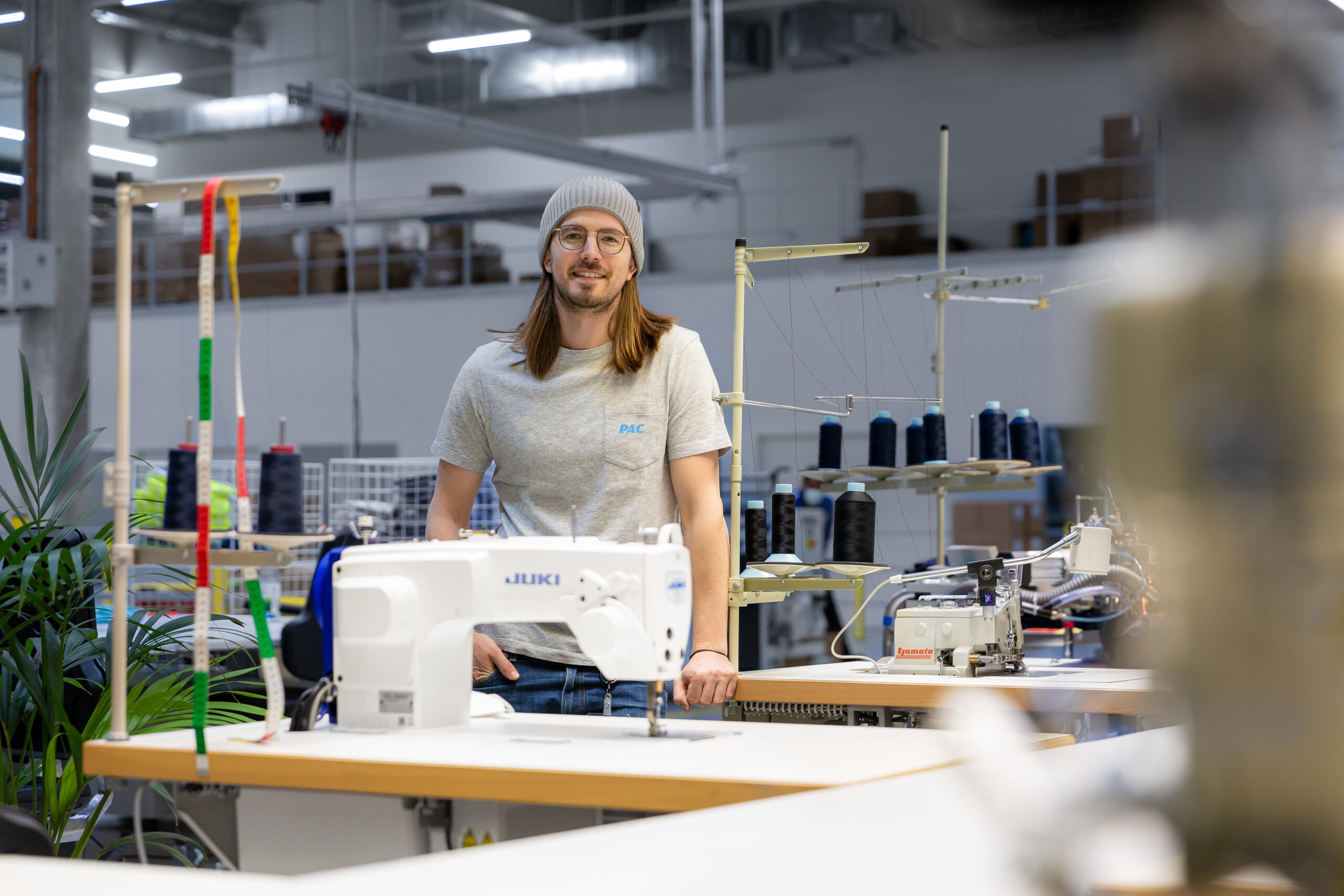 Mann mit Mütze steht in einer modernen Ötztaler Näherei mit Maschinen und Garnspulen.
