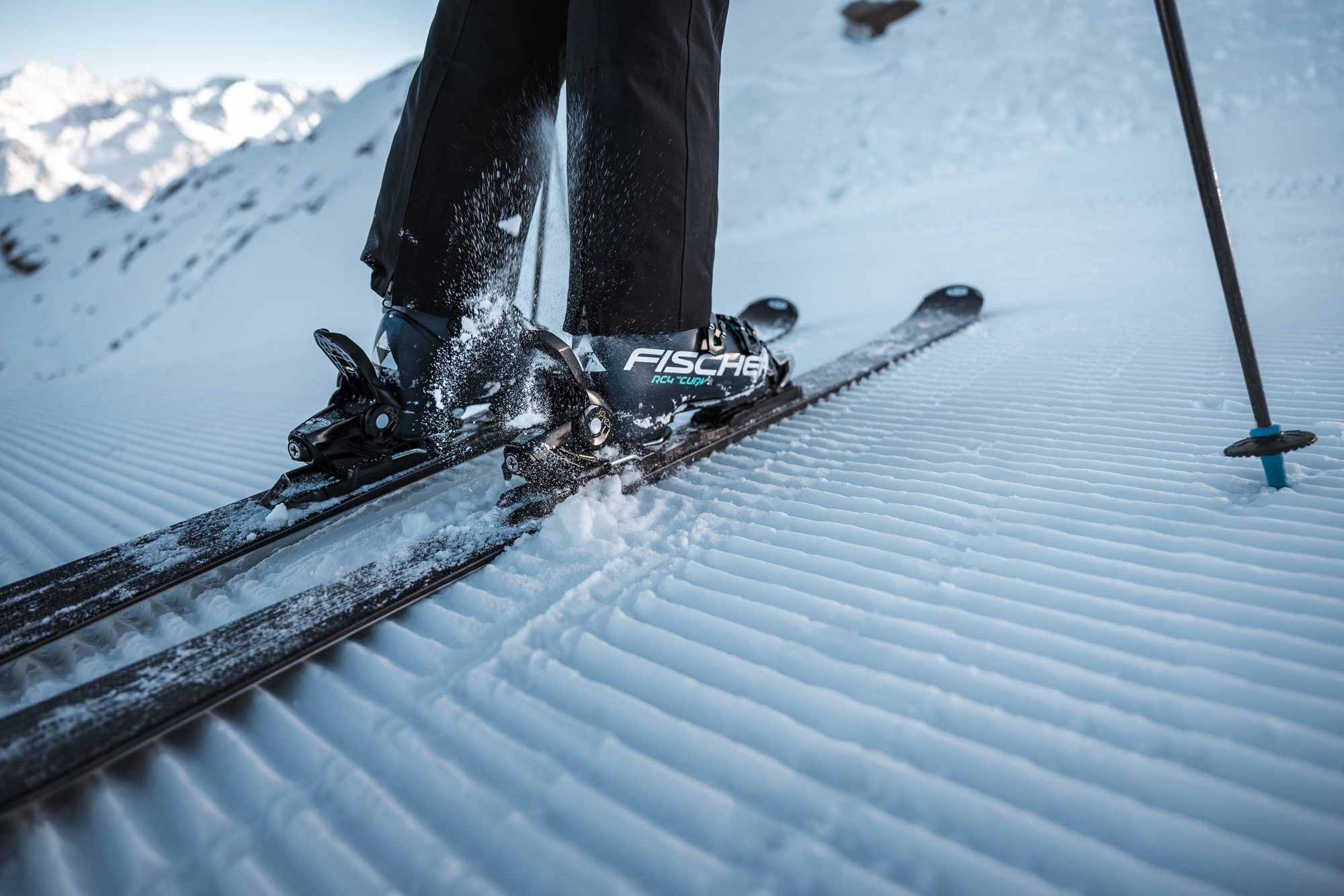 Nahaufnahme von Skischuhen, die in die Skier geschnallt sind, auf dem frisch präparierten Schnee des Ötztals mit den Bergen im Hintergrund.