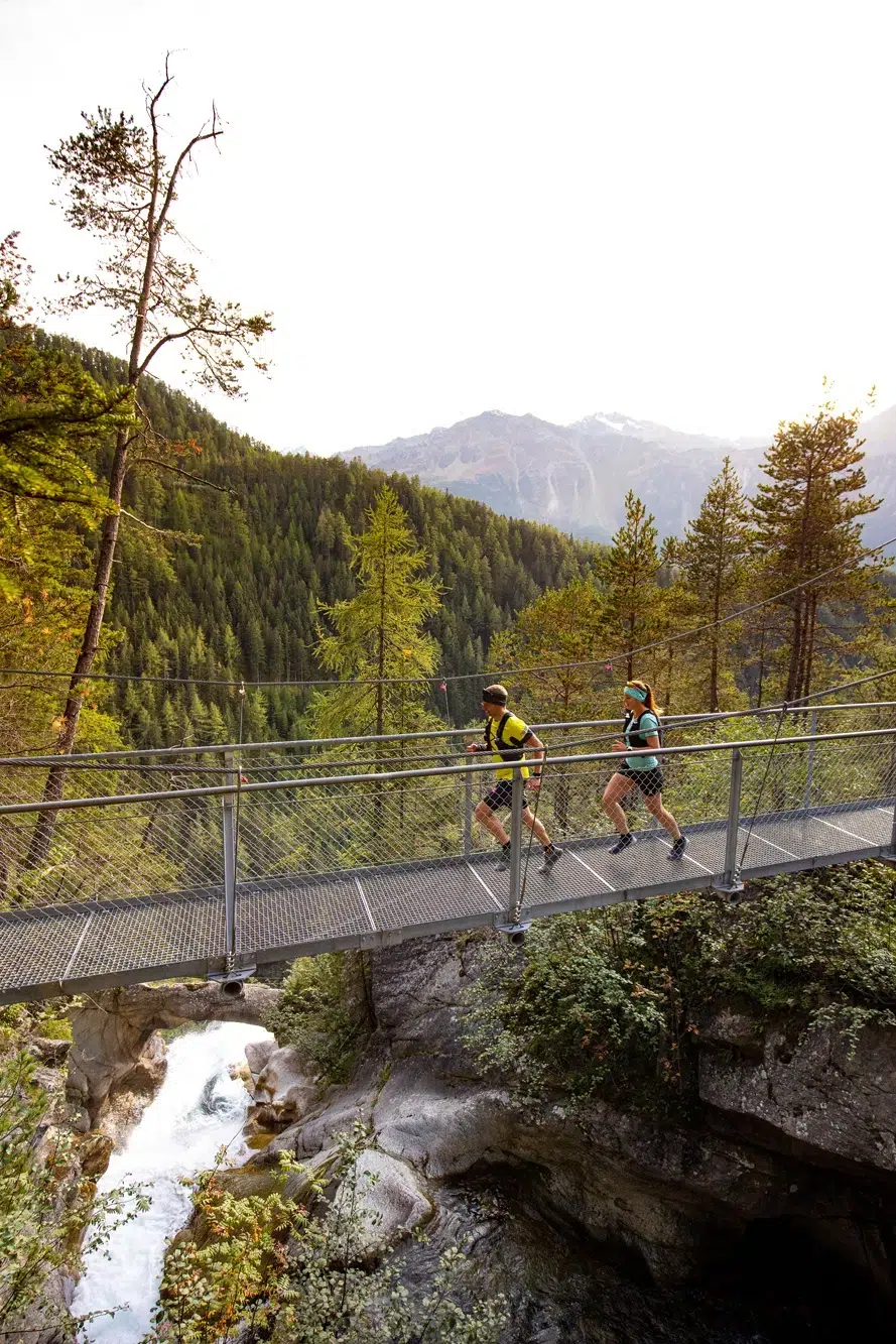 Zwei Personen laufen über eine Metallbrücke über ein bewaldetes Ötztal mit Bergen dahinter.