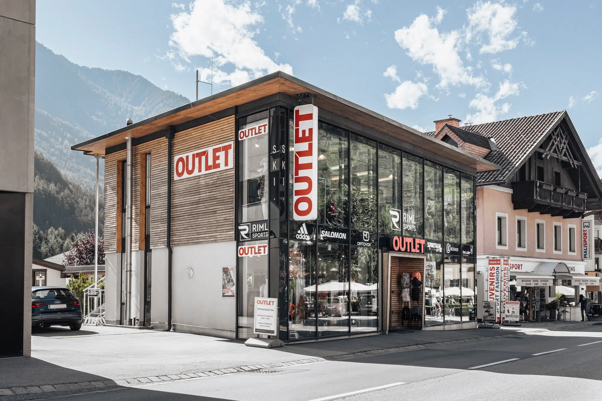 Modernes gläsernes Outlet-Geschäft mit großen OUTLET-Schildern, an einer sonnigen Ötztaler Straße mit den Bergen im Hintergrund gelegen.