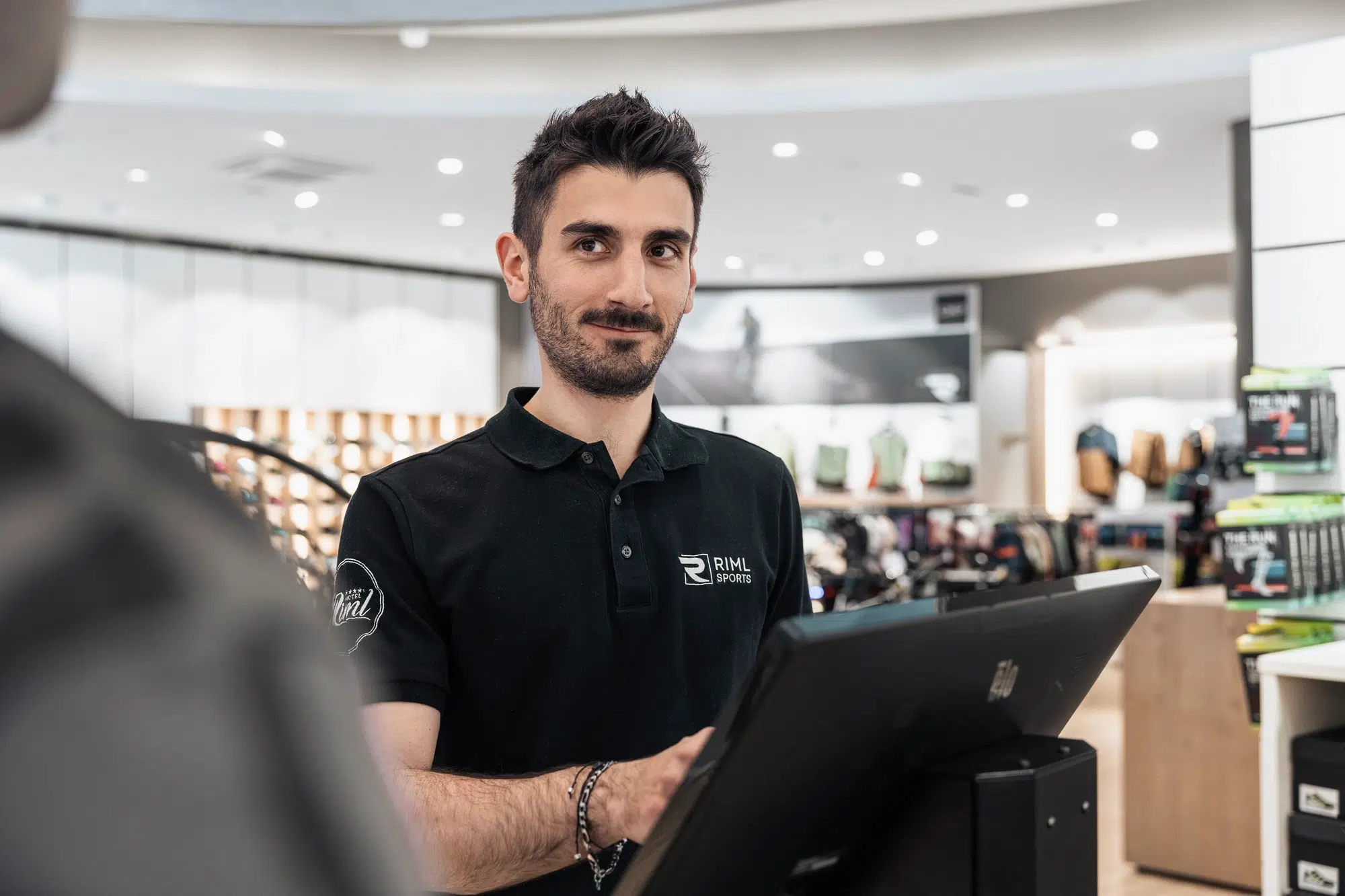 Ein Mann in einem schwarzen Polohemd steht in einem Ötztaler Geschäft hinter einem Tresen und arbeitet an einem Touchscreen.