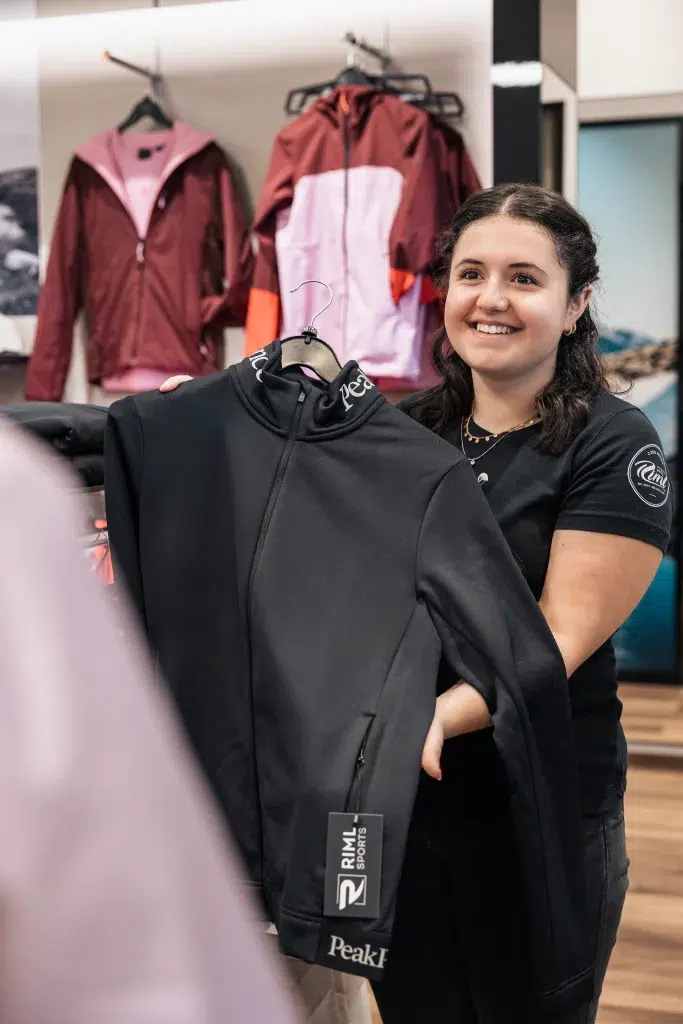 Lächelnde Frau mit einer schwarzen Peak Performance Jacke in einem Ötztaler Bekleidungsgeschäft.
