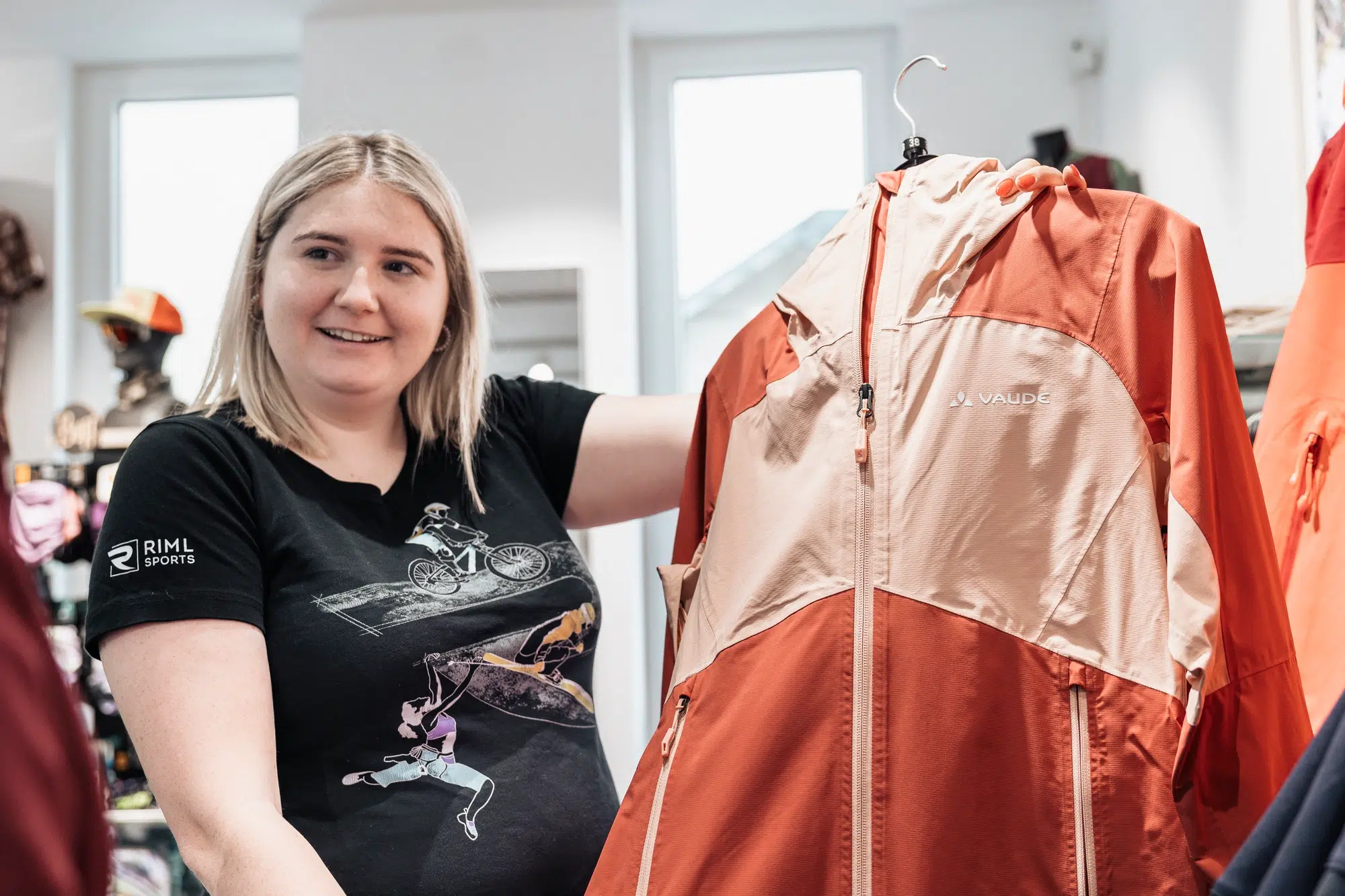 Eine Frau lächelt, während sie eine orange-beigefarbene Ötztaler Jacke in einem Bekleidungsgeschäft hochhält.