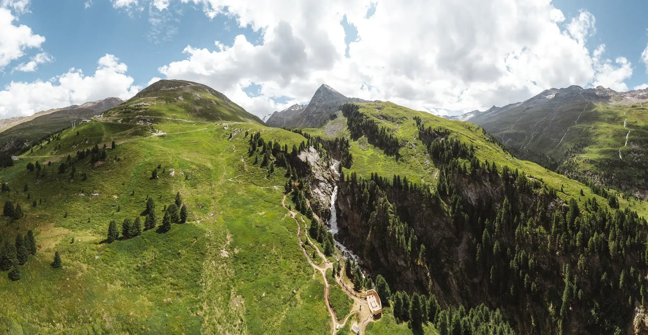 Luftaufnahme der grünen Ötztaler Berge mit einem Wasserfall, Bäumen und einem bewölkten Himmel.
