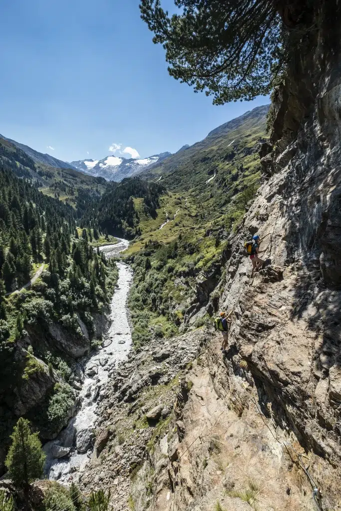 Zwei Wanderer erklimmen einen steilen felsigen Berg im Ötztal über einem Flusstal und fernen Gipfeln.