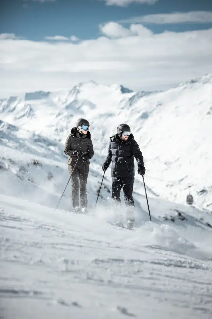 Zwei Personen beim Skifahren auf einem Ötztaler Berghang mit verschneiten Gipfeln im Hintergrund.