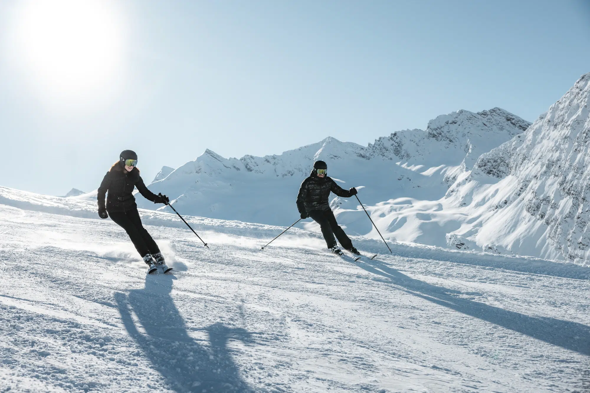 Zwei Personen fahren bei klarem, sonnigem Himmel einen verschneiten Ötztaler Berghang hinunter.