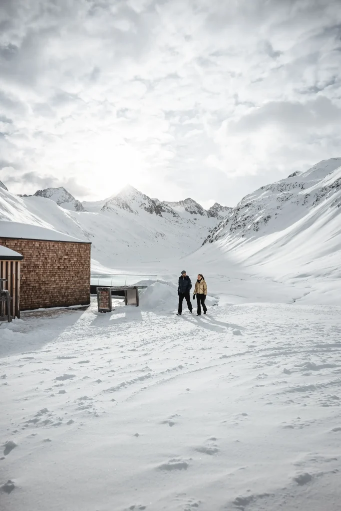 Zwei Personen gehen bei bewölktem Himmel in den verschneiten Ötztaler Bergen auf Schnee in der Nähe eines Holzhauses.