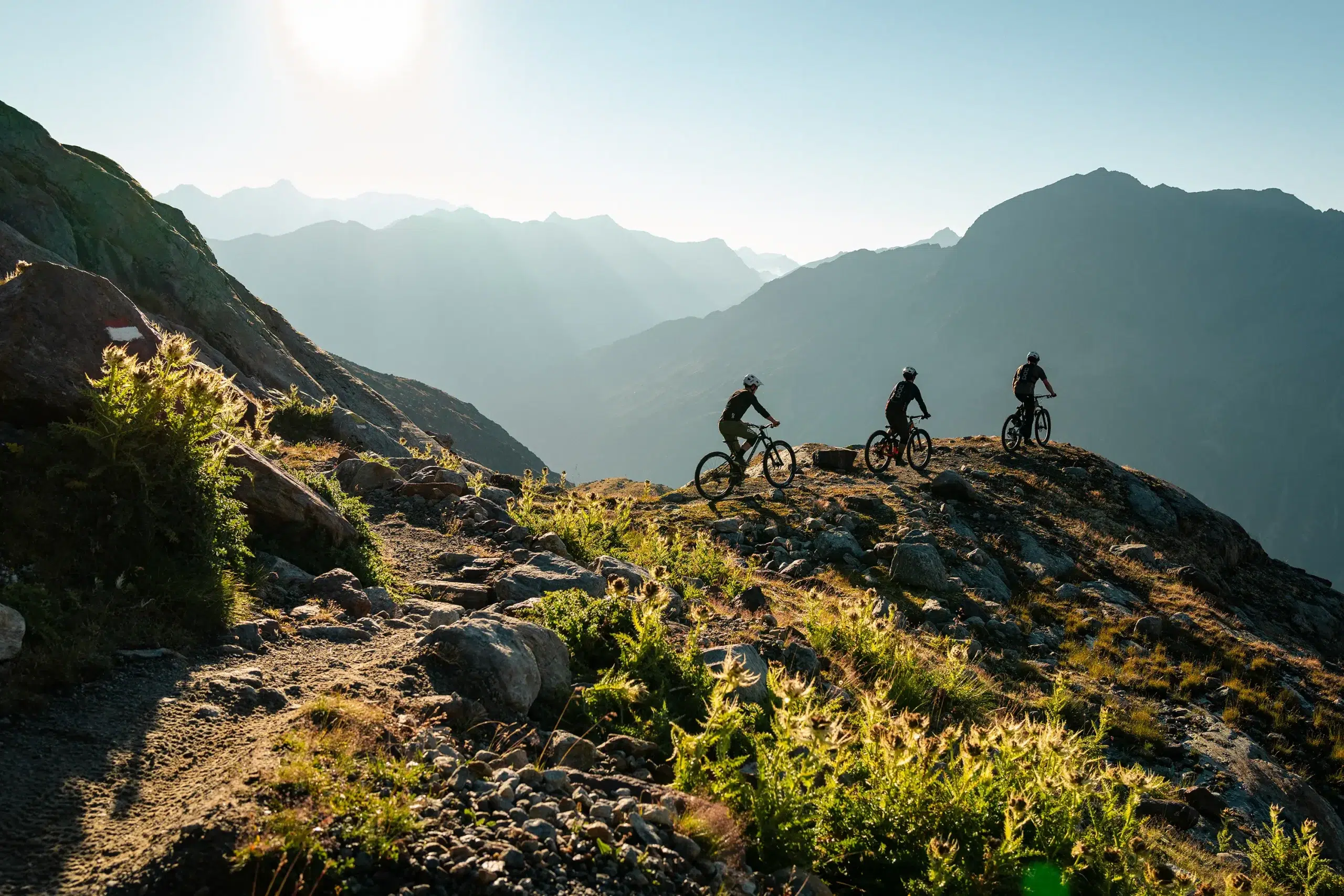 Drei Personen fahren mit dem Mountainbike auf einem felsigen Ötztaler Weg mit Bergen und Sonnenlicht im Hintergrund.