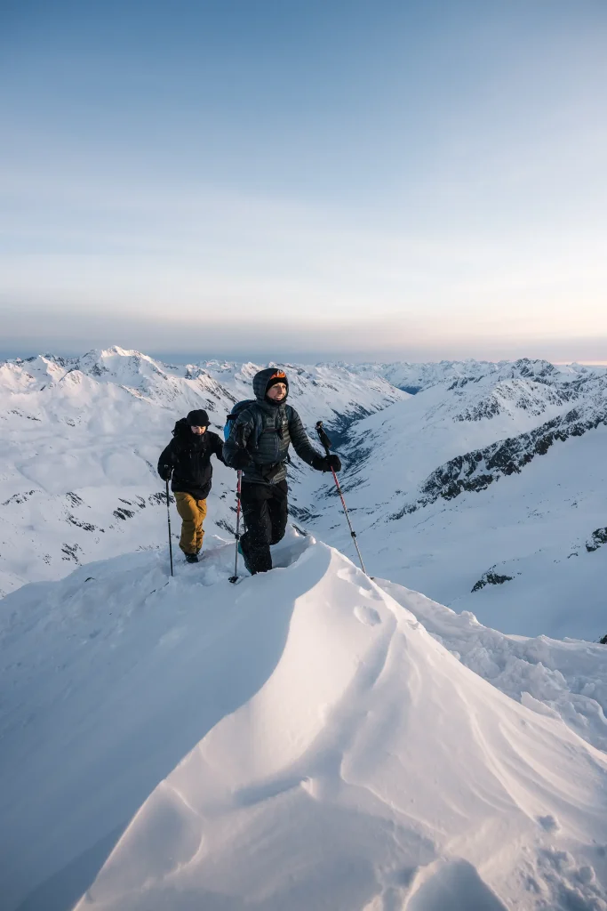 Zwei Personen wandern auf einem verschneiten Ötztaler Bergrücken, hinter ihnen ferne schneebedeckte Gipfel.