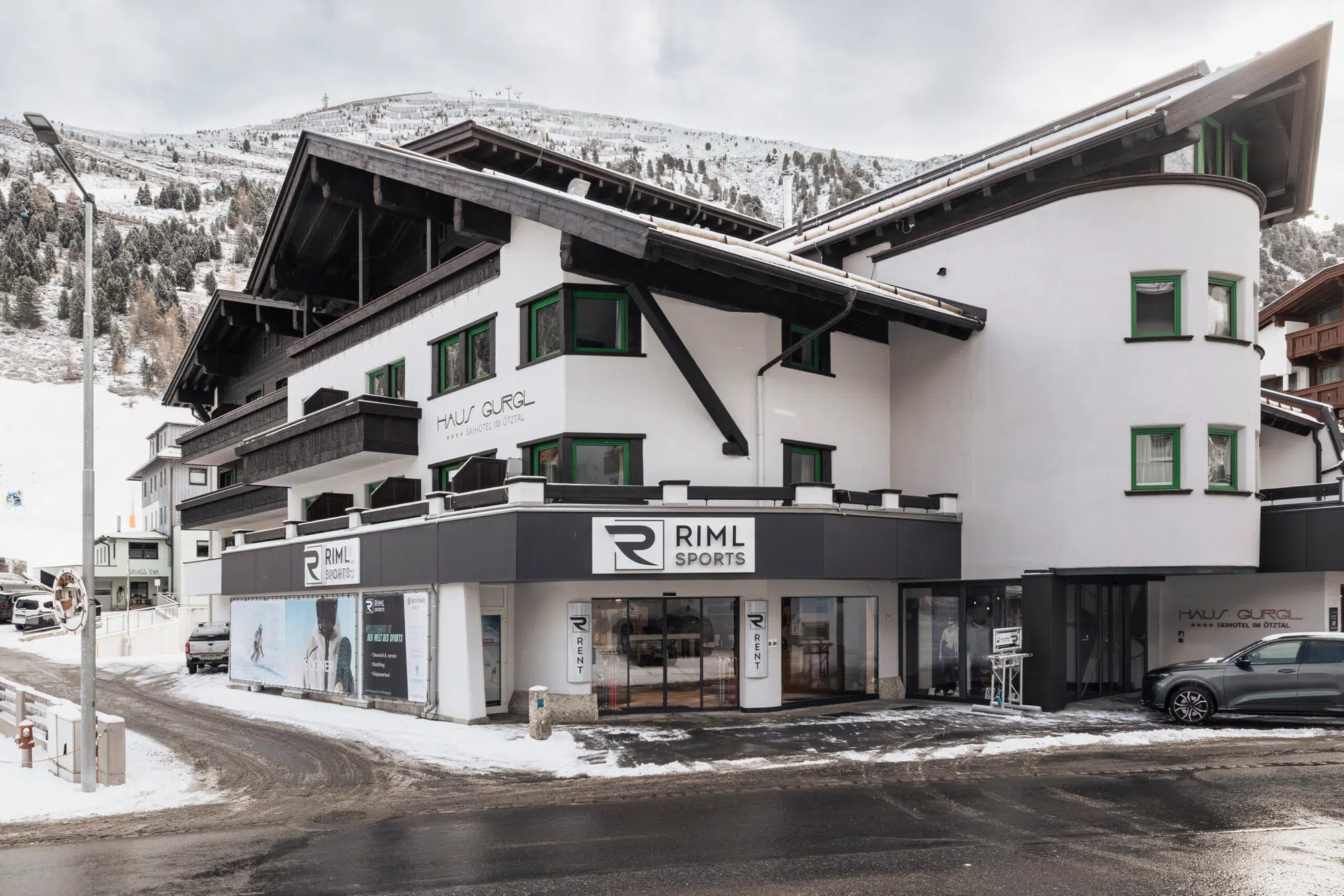 Weißes alpines Gebäude mit grünen Fenstern in einer verschneiten Bergstadt, RIML SPORTS-Schild sichtbar.