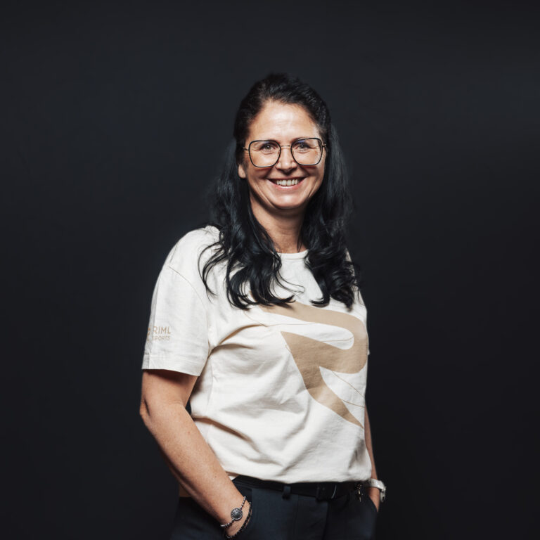 Frau mit langen dunklen Haaren und Brille, lächelnd, Teil unseres Jobs & Teams, auf dunklem Hintergrund.