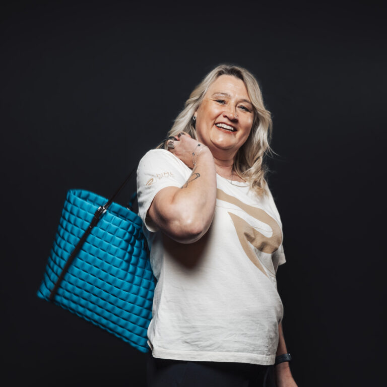 Lächelnde Frau mit einer blauen Tragetasche, bereit für neue Jobs & Teamchancen, vor einem dunklen Hintergrund.