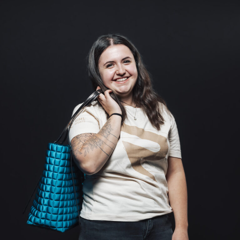 Lächelnde Frau mit langen Haaren, blaue Tasche über der Schulter, steht vor schwarzem Hintergrund-Jobs & Team.