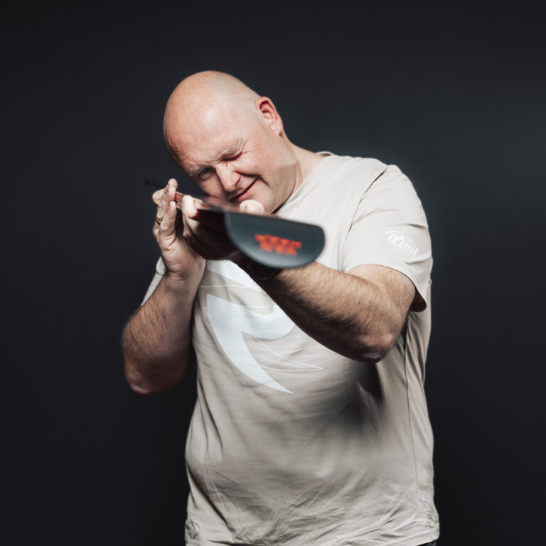 Mann mit Glatze und beigem Hemd, der mit einem Paddel nach unten zielt, vor einem dunklen Hintergrund.