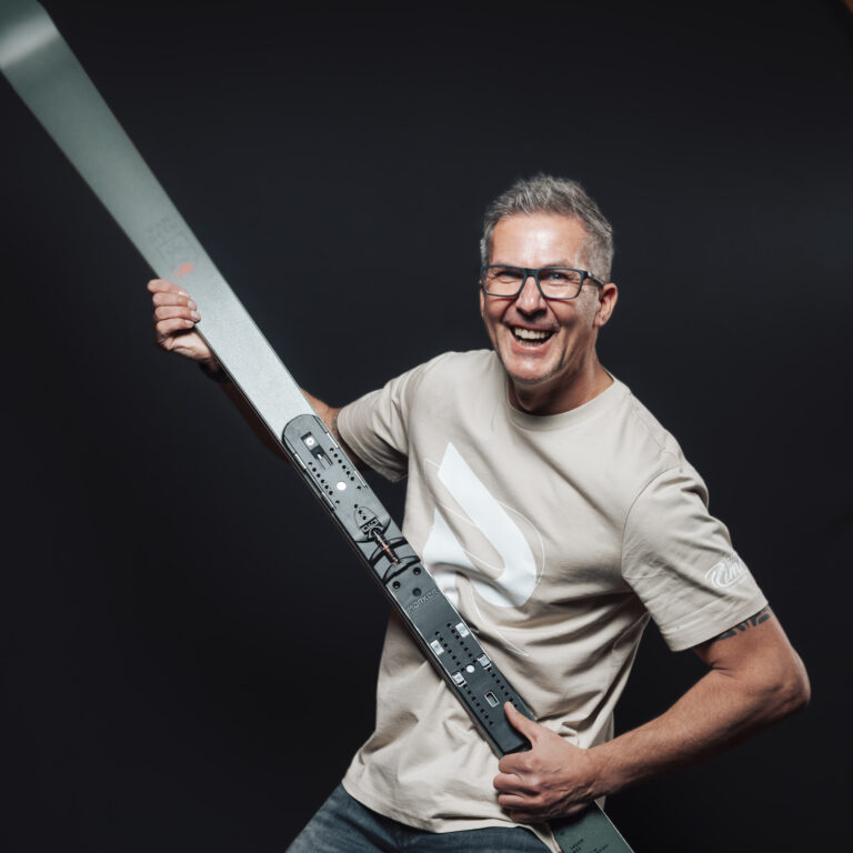 Lächelnder Mann mit Brille, Teil unseres Jobs & Teams, hält einen großen Ski vor einem dunklen Hintergrund.