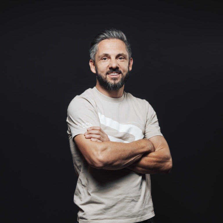 Mann mit Bart und grauem Haar, lächelnd, verkörpert Jobs & Teamgeist, Arme verschränkt vor dunklem Hintergrund.