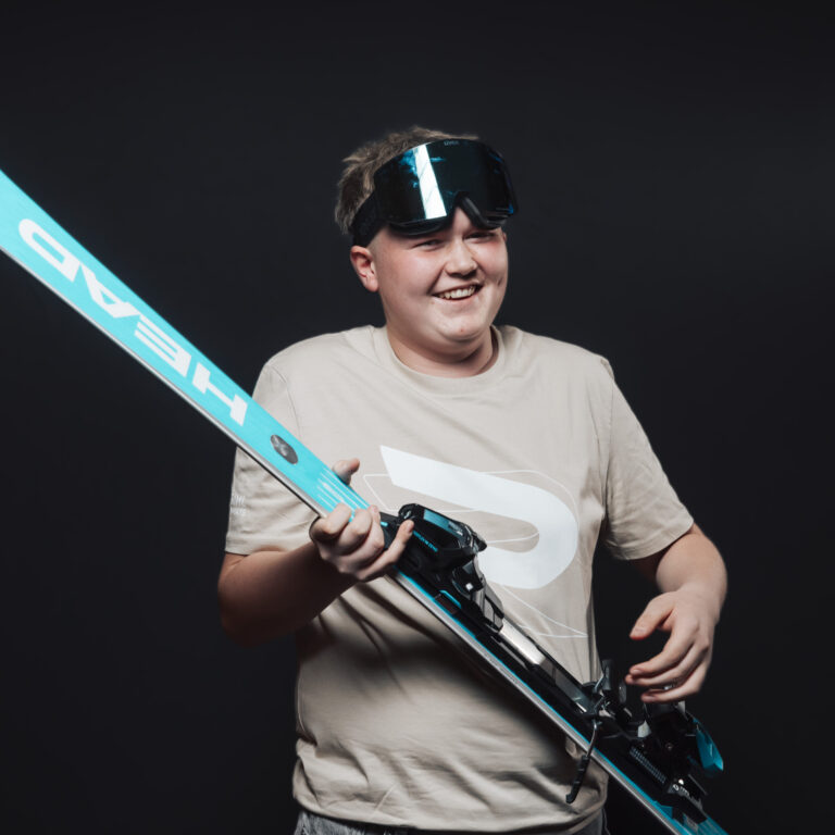 Lächelnde Person, die blaue Skier hält und eine Skibrille vor einem dunklen Hintergrund trägt.