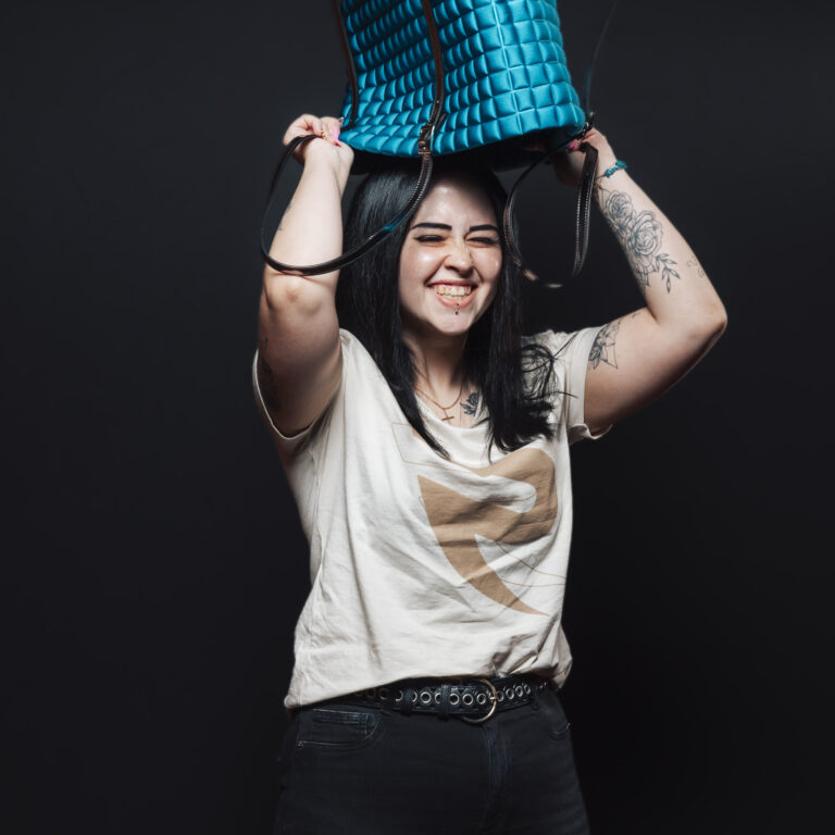 Eine lächelnde Frau, die sich spielerisch eine blaue Tasche über den Kopf hält und sich über den Job und den Teamgeist freut.