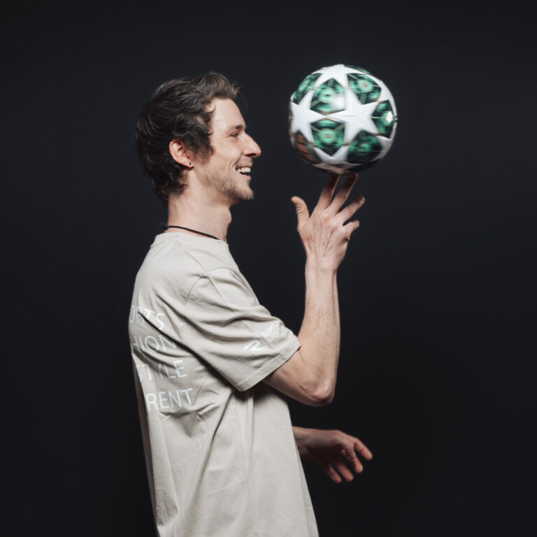 Ein lächelnder Mann, der einen grün-weißen Fußball dreht, verkörpert Teamgeist vor einem schwarzen Hintergrund.