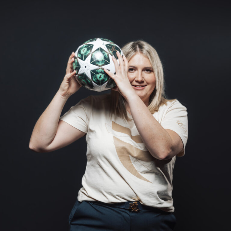 Lächelnde Frau mit einem Fußball in der Hand, die Teamarbeit und Teamgeist vor einem dunklen Hintergrund hervorhebt.