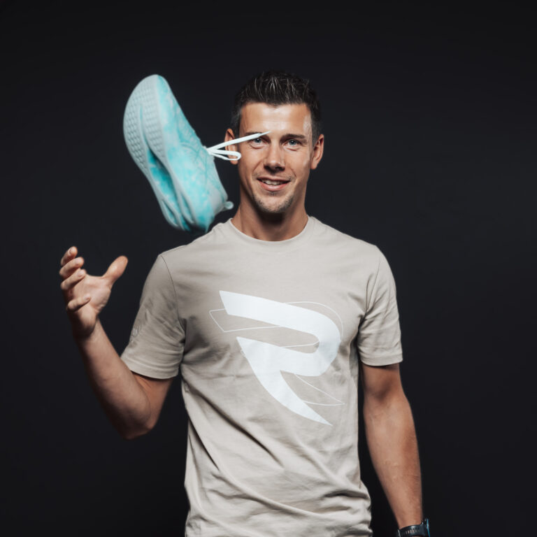 Ein lächelnder Mann wirft einen blauen Turnschuh, der Jobs & Team repräsentiert, vor einem schwarzen Hintergrund.