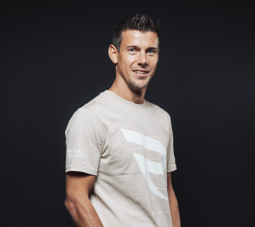 Ein Mann in einem beigen T-Shirt mit einem großen weißen Logo, das Jobs & Team repräsentiert, steht vor einem schwarzen Hintergrund.