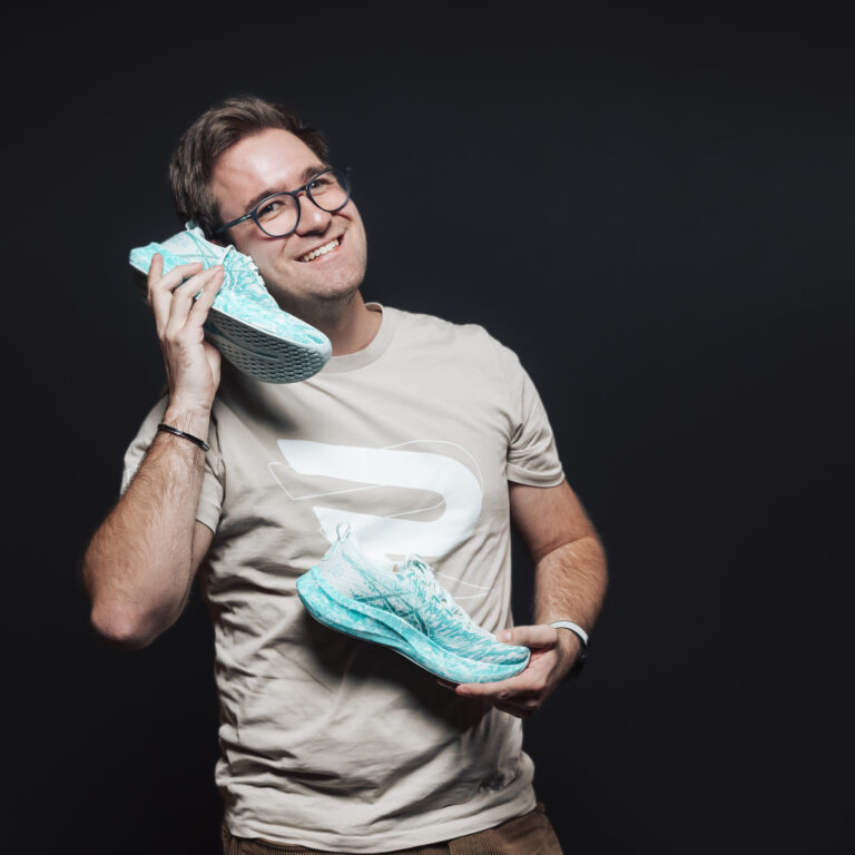 Ein lächelnder Mann hält blaue Turnschuhe, einen bis zum Ohr, und zeigt damit Jobs & Teamgeist.