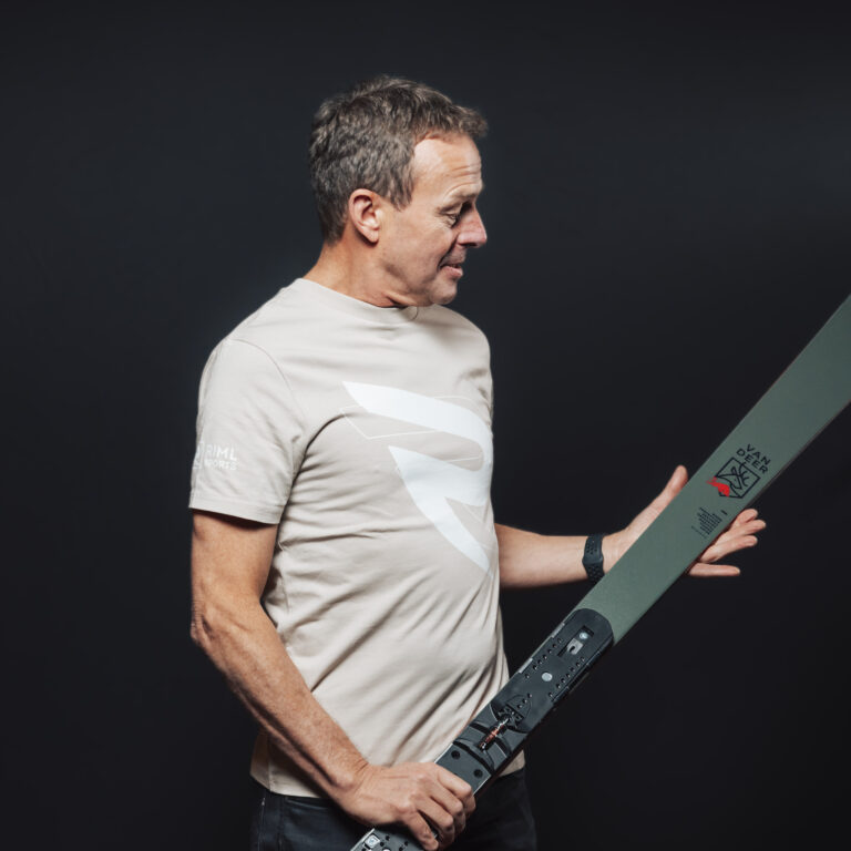 Mann in beigem T-Shirt, der einen Ski hält, als Symbol für unsere Jobs & Teamgeist vor schwarzem Hintergrund.