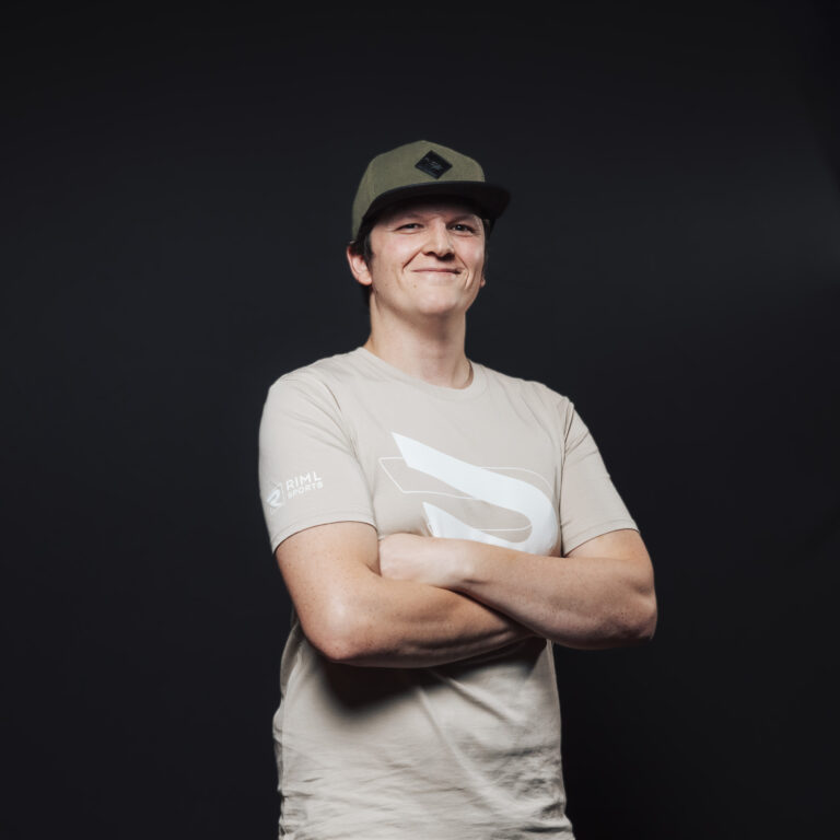 Eine Person im hellbraunen Hemd und mit grüner Mütze steht mit verschränkten Armen da und verkörpert Teamgeist.