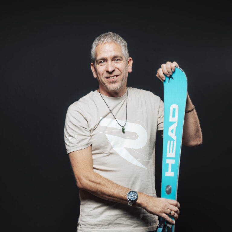 Ein lächelnder Mann von Jobs & Team hält einen blauen HEAD-Ski vor einem dunklen Hintergrund.