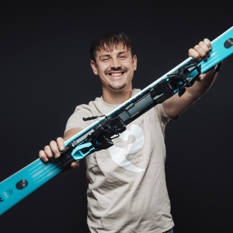 Lächelnder Mann, Teil unseres Jobs & Teams, hält blaue Skier vor einem schwarzen Hintergrund.