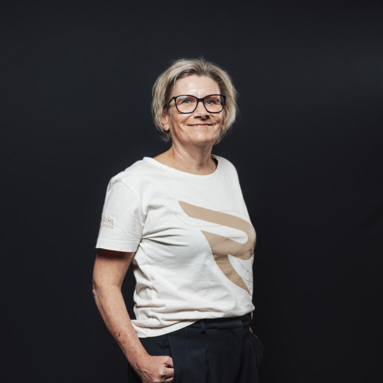 Eine lächelnde ältere Frau mit Brille, kurzen Haaren und weißem T-Shirt repräsentiert Jobs & Team.