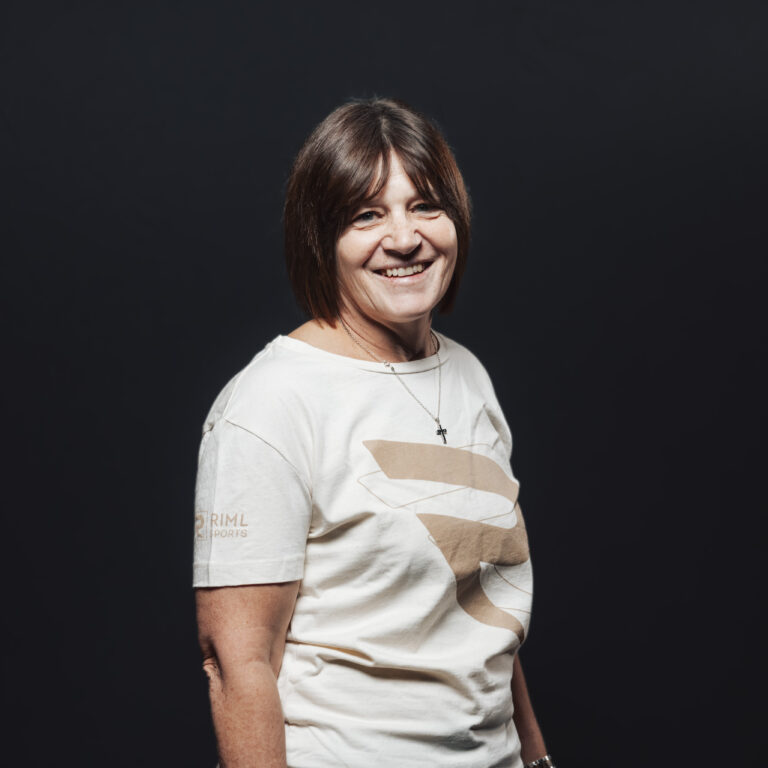 Frau mit kurzen braunen Haaren lächelnd, Teil unseres Jobs & Teams, vor einem dunklen Hintergrund.