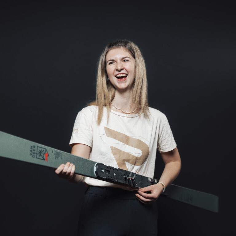 Lächelnde Frau, die einen Ski in der Hand hält, als Symbol für unsere Jobs & Teamgeist, vor einem dunklen Hintergrund.