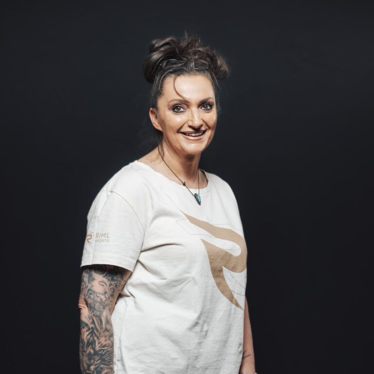 Lächelnde Frau mit Tattoos in einem weißen Hemd posiert, die Jobs und Teamgeist repräsentiert, vor dunklem Hintergrund.