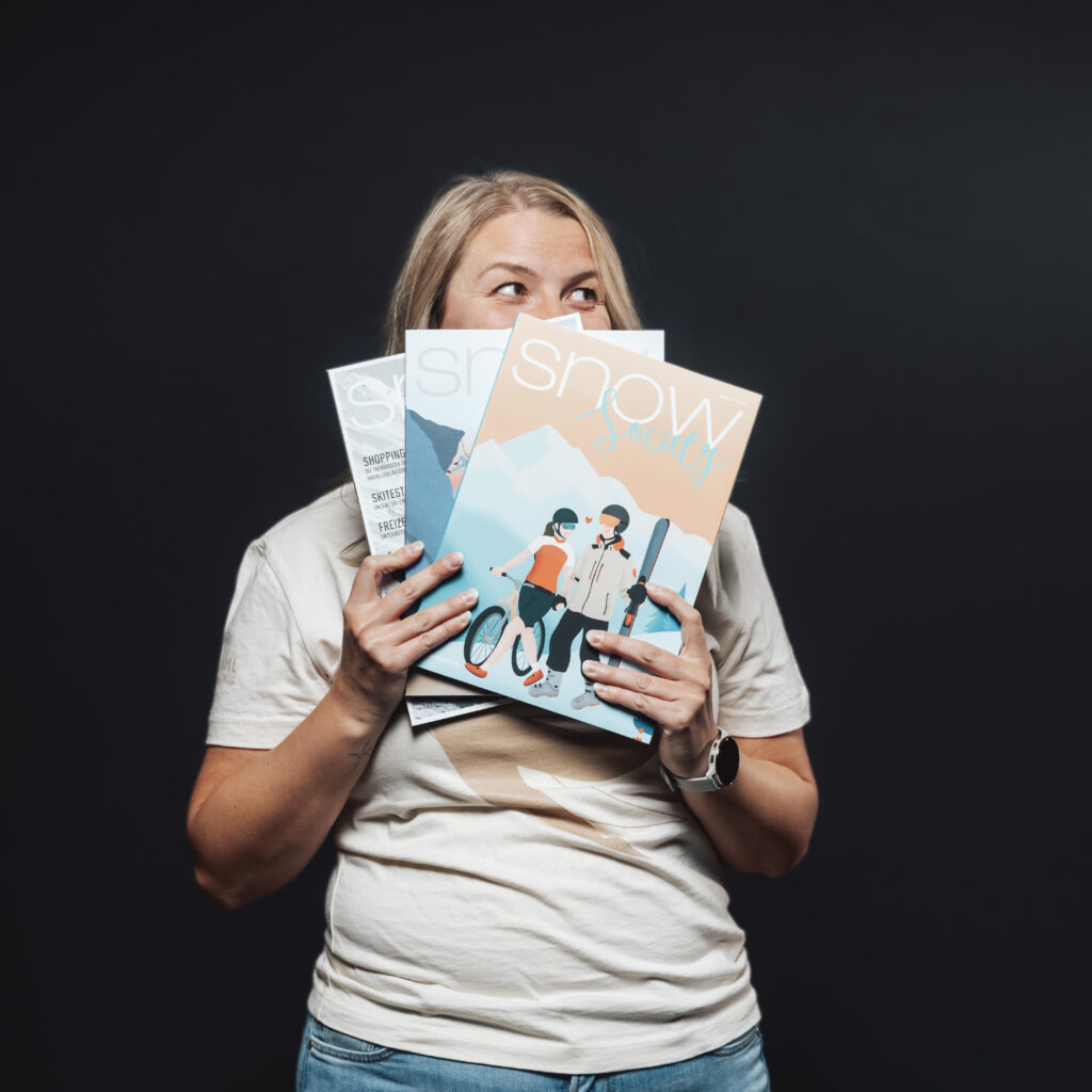 A woman holds up three Snow Society magazines—exploring Jobs & Team—against a dark background.